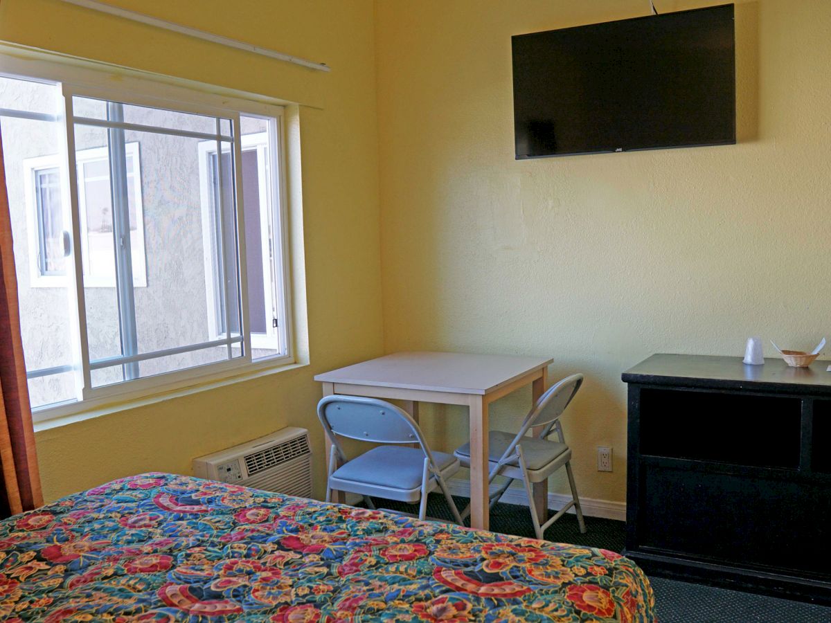 A small room with a patterned bed, table and two chairs, TV on wall, window, curtain, and a cabinet with some items.