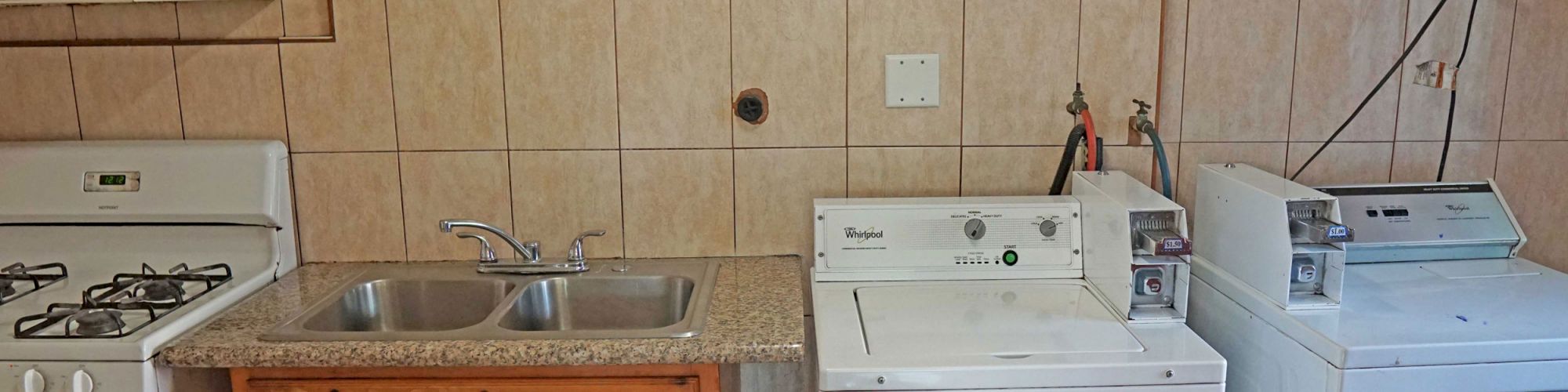 The image shows a kitchen/laundry area with a stove, sink with a wooden cabinet, a washing machine, and a dryer on a tiled wall.