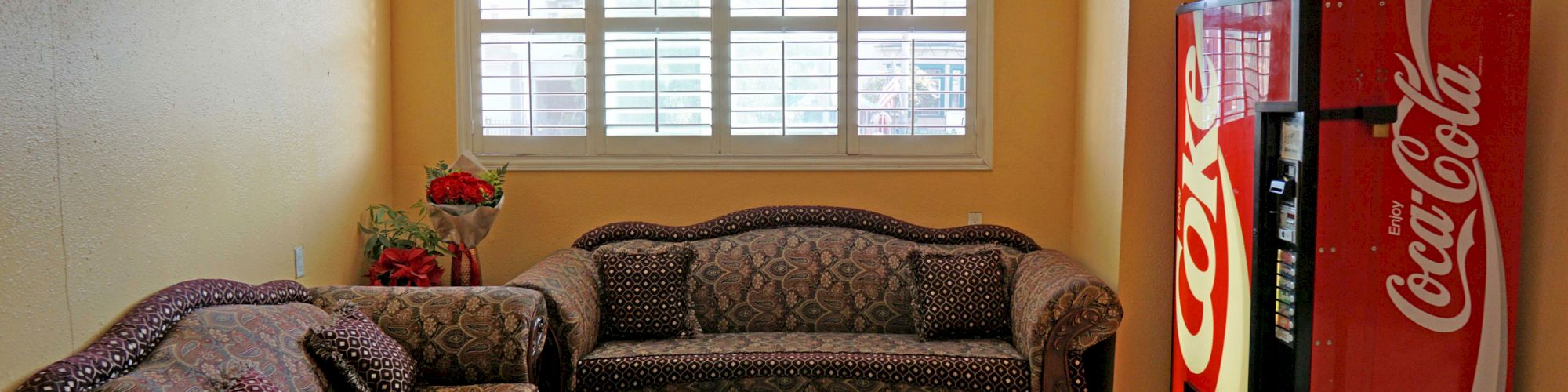 The image shows a small room with patterned sofas, a coffee table, a flower vase, and a Coca-Cola vending machine. The walls are painted yellow.