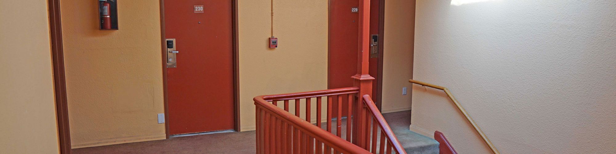 A hallway with two orange doors, a staircase, an exit sign, a fire alarm, and a bright light from a window on the wall.