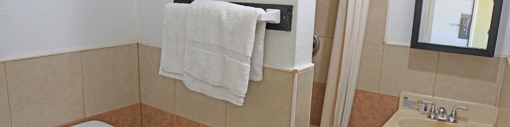 The image shows a small bathroom with a toilet, a shower with a beige curtain, a sink with a mirror, and a towel hanging on a rack.