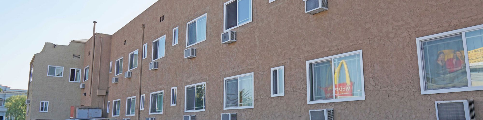 Image of a beige hotel building with multiple windows and air conditioning units, with a white car parked in front.