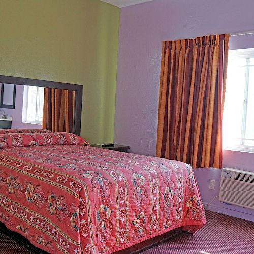 A bedroom with a large bed covered in a red floral bedspread, a window with orange curtains, a mirror above the bed, and an air conditioner.