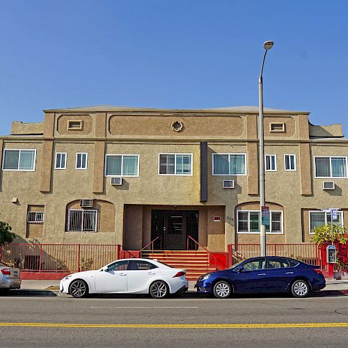 An apartment building with three cars parked in front on the street. The building has a beige exterior and is adorned with several windows.