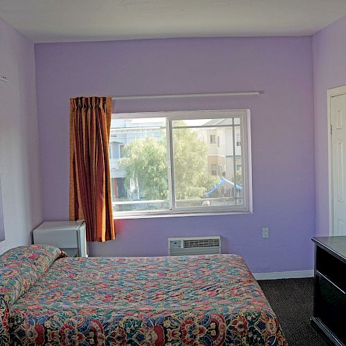 The image shows a hotel room with light purple walls, a bed, a window, a TV mounted on the wall, a small fridge, and a dresser.