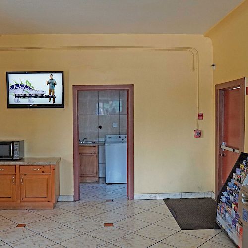 The image depicts a small room with a microwave on a cabinet, a wall-mounted TV, a door leading to a laundry area, and an ATM or vending machine.