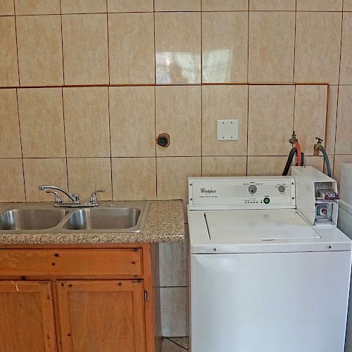 The image shows a kitchen and laundry area with a stove, a double sink, a washing machine, and a dryer, all set against a tiled wall.