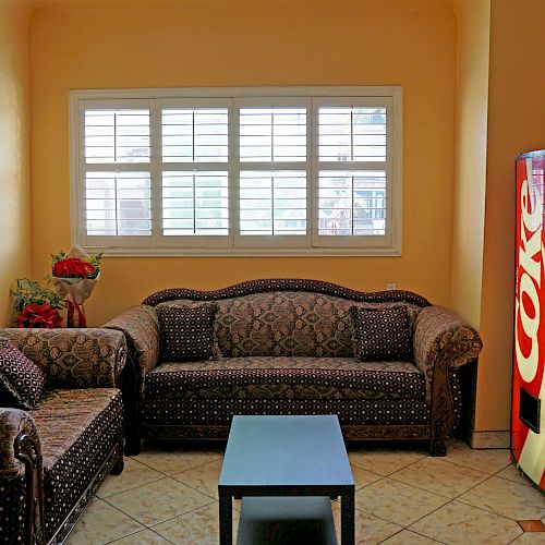 A cozy room with patterned couches, a coffee table, a window with blinds, a Coca-Cola vending machine, and some flowers on the couch.