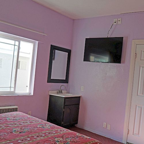 A bedroom with purple walls, a bed with a patterned red cover, TV mounted on the wall, sink with mirror, window with curtains, and a white door.