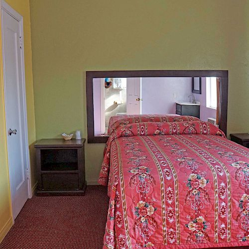 This image shows a small bedroom with a bed featuring a bright floral bedspread, a mirror behind the bed, and a mini-fridge in the corner.