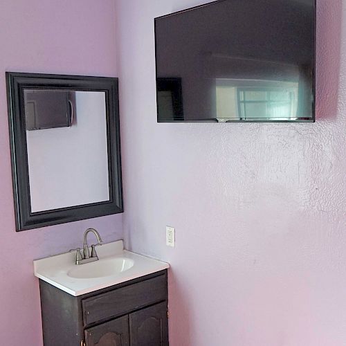 The image shows a purple wall with a mounted TV, a wall-mounted sink with a mirror above it, and an electrical socket beside the mirror.