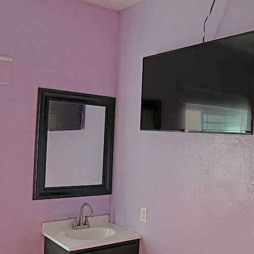 The image shows a room with lavender walls, featuring a wall-mounted TV, a mirror above a sink with a faucet, and a window on the left side.