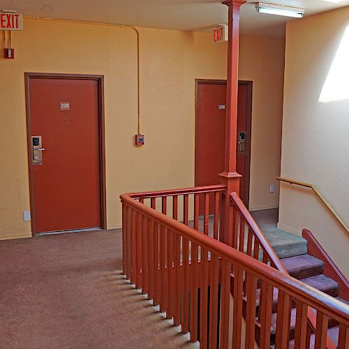 The image shows an indoor hallway with two red doors, a staircase, a fire alarm, and an exit sign. The walls are light-colored.