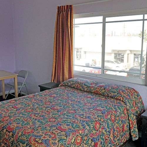 The image shows a simple room with a bed covered in a colorful patterned bedspread, a small table with two chairs, and two large windows with curtains.