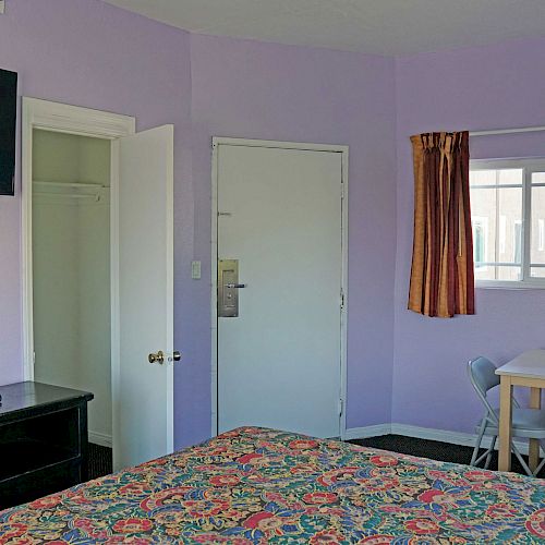 A room with lavender walls, a bed, TV, open closet, table with chairs, door, and window with orange curtains. Ends the sentence.