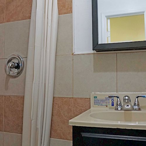 A bathroom with a tiled shower, a curtain, a sink with a mirror above it, and a small cabinet below the sink.