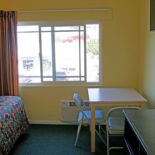 A small room with a bed, table with chairs, dresser, wall-mounted TV, window with curtain, and simple decor in a tidy arrangement.