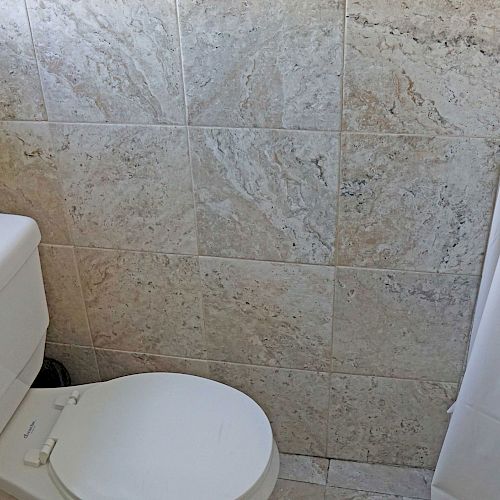 The image shows a small bathroom with beige tile walls, a white toilet, and a white shower curtain. The space appears clean and simple.