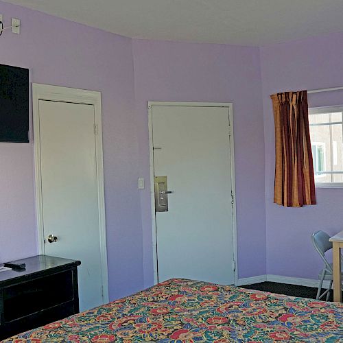 The image shows a simple hotel room with light purple walls, a bed with colorful bedding, a wall-mounted TV, a small table with a chair, and a window.