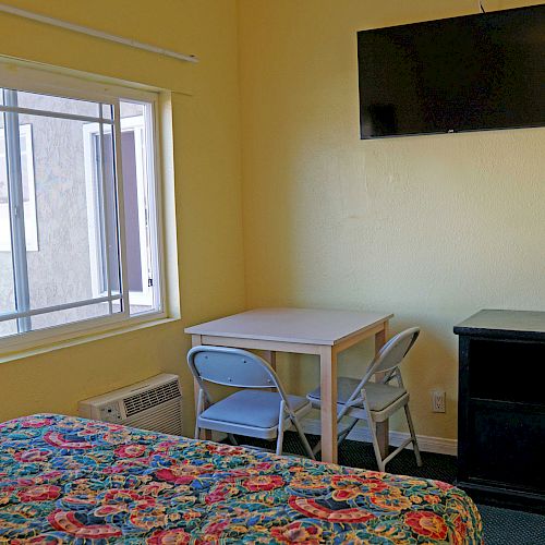 A small room with a bed, a window, a table with two folding chairs, a wall-mounted TV, and a dresser with some items on it.