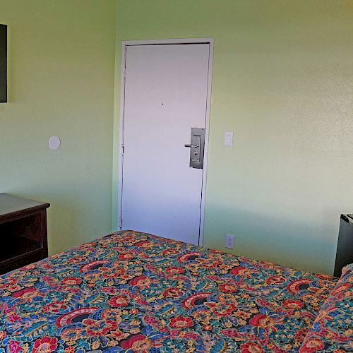 A room with a bed, colorful bedspread, TV, door, dresser, and mini-fridge against green walls, well lit with natural light.