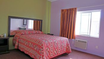 The image shows a bedroom with light-colored walls, a bed with a floral red bedspread, an orange curtain, a window, and an air conditioner unit.