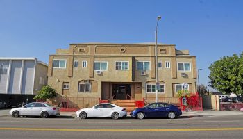 The image shows a two-story building with a beige facade, located on a street with three parked cars and a tree on the right side.