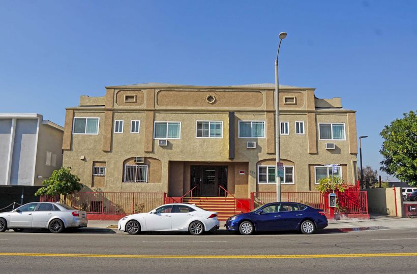 The image shows a two-story building with a beige facade, located on a street with three parked cars and a tree on the right side.