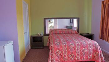 A small bedroom with a bed covered in a floral-patterned quilt, a nightstand, a mini-fridge, and a large mirror over the headboard.