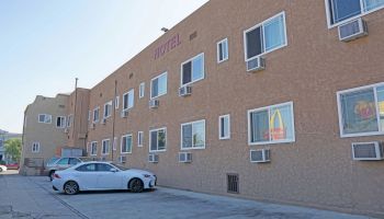 An exterior view of a two-story motel building with multiple windows and air conditioning units, featuring parked cars in the lot.