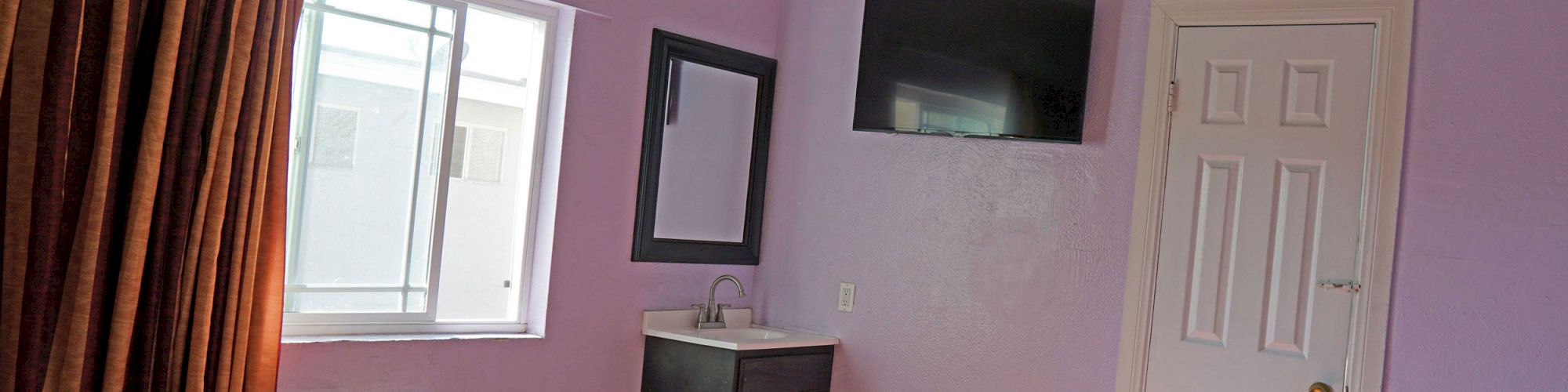 The image shows a hotel room with a purple wall, a bed with a red patterned cover, a window, a black TV mounted on the wall, and a small sink area.