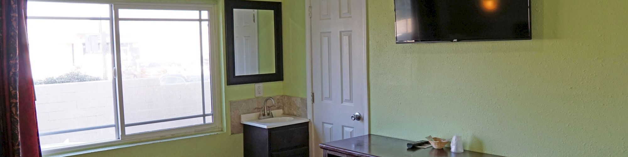 A small room with a TV on the wall, bed with colorful quilt, window, dresser, bathroom vanity, and green walls.