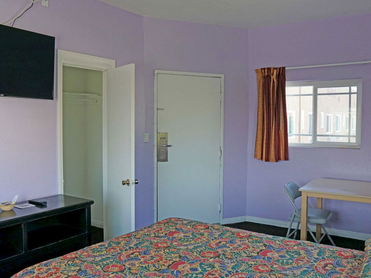 A room with purple walls featuring a TV, a door, a window with curtains, a small table with chairs, bed with patterned cover, and some furnishings.