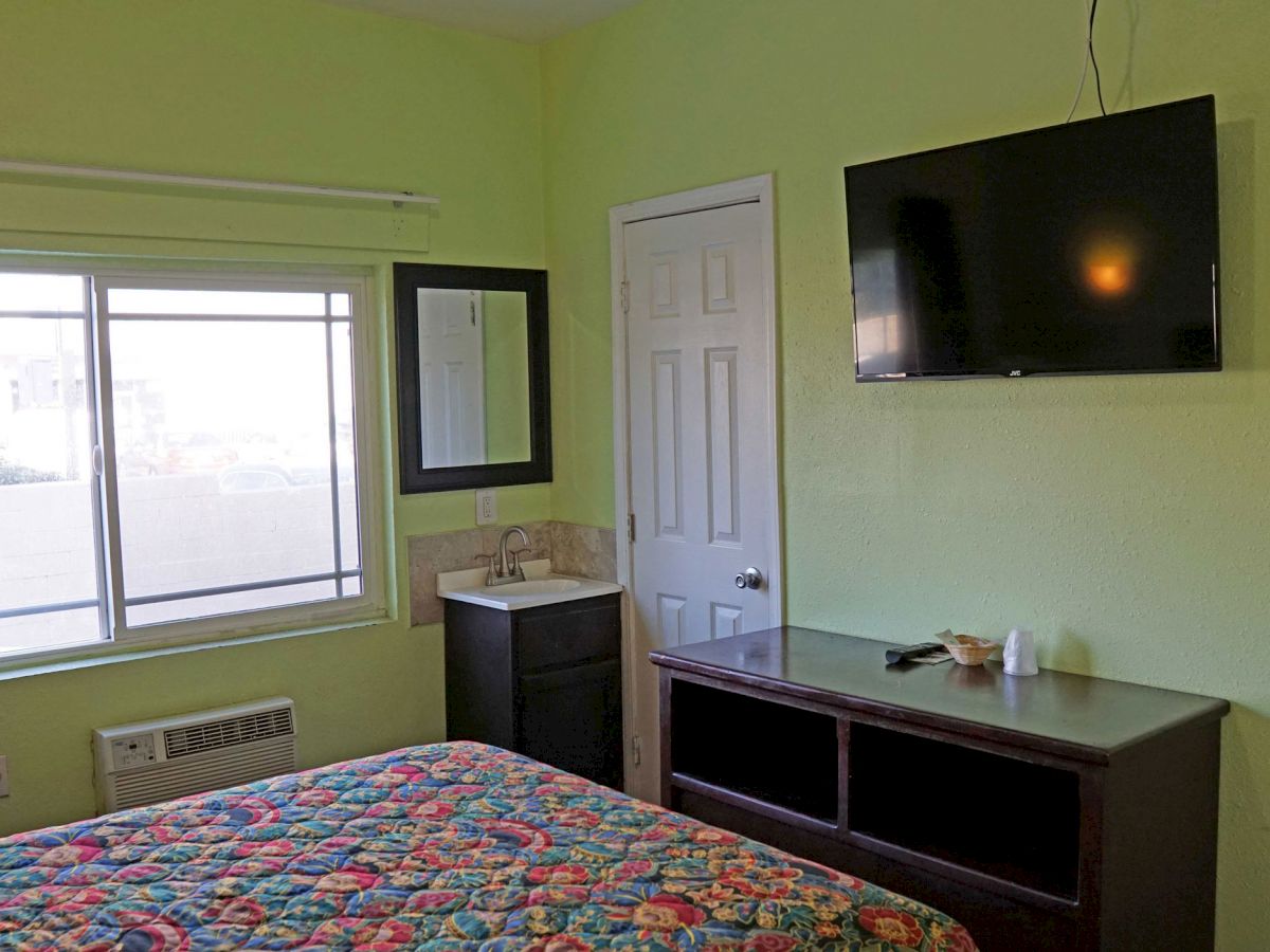 The image shows a small hotel room with a green wall, a bed with a colorful quilt, a wall-mounted TV, a window, and a small sink area.